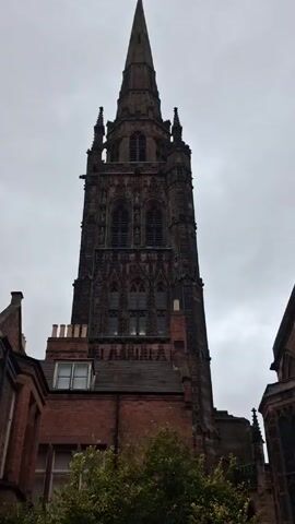 The video shows a tall, dark stone church tower with a pointed spire against a cloudy sky. The tower is intricately designed with multiple windows and spires. A red brick building with a gabled roof and chimneys is in the foreground. A green bush and a green container sit in front of the building.  There is also a red trash can in the scene. The video is taken from a low angle, emphasizing the height of the church tower.