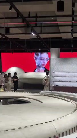 The video depicts a spacious, modern retail or exhibition interior with a high, dark ceiling and track lighting. A large, curved, multi-tiered, light-grey structure, resembling a concrete track, dominates the foreground and midground. Several large, smooth, matte-grey, spherical pods or capsule-like objects are moving continuously from left to right along this track; one pod is clearly visible with three circular indentations on its side. In the background, a large LED screen displays a vibrant red background with a stylized close-up of a person's head wearing futuristic, chrome-like eyewear. To the right of the screen, grey textured walls feature shelves displaying various eyeglasses or sunglasses. Approximately ten individuals are present: a group of four women in light-colored clothing are standing together on the left side of the track, observing the moving pods. A man in dark clothing stands near them, holding a dark object, seemingly overseeing the operation of the display. Two other women are observing from the right side of the track, and additional individuals are visible near the eyewear shelves in the background.