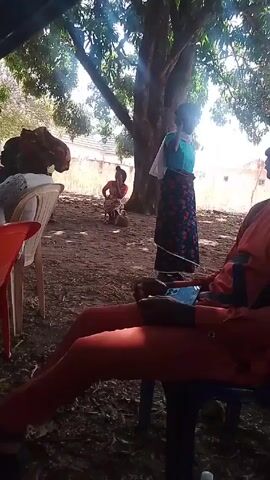 The scene is an outdoor gathering with individuals in traditional African clothing. A person in a red outfit is seated on a blue plastic chair, focused on their phone. In the background, other individuals are walking and milling around under the shade of a large tree. There is a building visible behind the tree in the background. The ground is dirt and scattered with leaves.