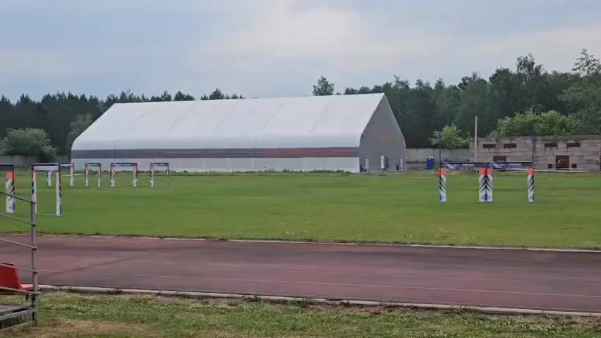 A drone flies through a course set up on a grassy field. The course consists of several tall, narrow gates made of white and red striped poles with blue and white flags. In the background, there is a large white tent-like structure and a smaller brick building with a chain-link fence. Trees line the far edge of the field. The drone appears to be controlled remotely and is maneuvering through the gates. The sky is overcast, suggesting a cloudy day.