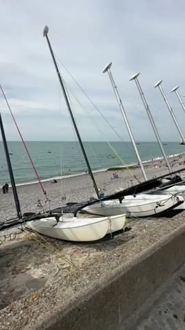 The video shows a view of a pebble beach with several small sailboats pulled up on the shore. The boats are white with tall masts. In the background, the sea is visible with a few people in the water and on the beach. The sky is cloudy with some patches of blue. The camera is positioned at a low angle, looking out towards the sea. There are also some concrete structures in the foreground.