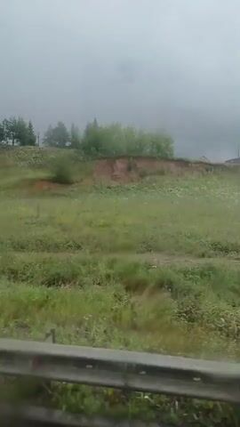 The video shows a landscape with a grassy hill on the right side of the screen and a guardrail in the foreground. A building with a blue roof is visible on top of the hill. The sky is overcast and it appears to be raining, as evidenced by the water droplets on the windshield. The overall impression is of a rural or suburban area.