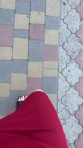 A first-person perspective video shows a person walking on a sidewalk. The sidewalk is paved with interlocking bricks in a pattern of red, yellow, and gray. To the right of the sidewalk, there is a decorative border of gray and pink interlocking bricks. The person is wearing a red dress and black sandals. The video then pans up to show a street scene. There are buildings on either side of the street, as well as trees and streetlights. The sky is clear and blue. Cars can be seen driving on the street in the distance.