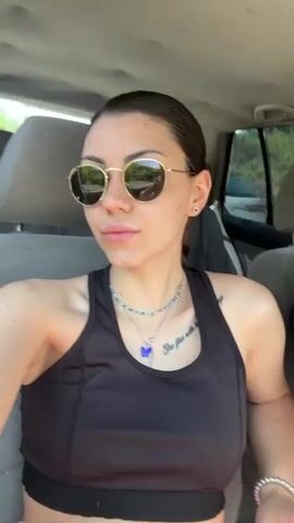 A young woman is seated in the front passenger seat of a car, illuminated by bright daylight. She has dark hair, slicked back into a low braid, and notable freckles across her nose and cheeks. Her eyes are concealed by gold-rimmed, round sunglasses that reflect the outdoor environment. She has a small stud piercing on her nose and multiple piercings on her ears.

She is wearing a black athletic crop top or sports bra, and several necklaces are visible: a beaded blue and green one, and two silver chains, one featuring a prominent blue butterfly charm. A white beaded bracelet adorns her right wrist. A tattoo on her upper right chest reads, "She flies with". The bottom edge of a pair of bright pink pants is visible at the very bottom of the frame.

Throughout the video, she looks directly at the camera with a relaxed and slightly smiling expression. Around the 0:02 mark, she briefly lifts her right hand, holding a small white object (possibly a hair tie or small stick), and runs her fingers through the end of her braid. The car's interior, including the light-colored seat fabric, is visible, and green foliage can be seen through the rear window, suggesting an outdoor location in Belopoptsi, Bulgaria. Background music is audible.