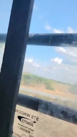 The video shows a view from inside a vehicle looking out through a dirty window. A dark, vertical bar is on the left side of the frame, and horizontal bars are in the foreground and background, obscuring the view. Through the window, a sunny, blue sky with scattered clouds can be seen. The landscape outside appears to be rural, with trees and vegetation visible. There is a road or pathway in the middle ground, and a yellow and black striped warning sign is visible on the left side of the frame at one point. The vehicle appears to be in motion, as evidenced by the shifting landscape. A sticker with text is partially visible on the lower part of the window.