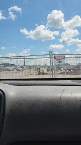 A view from inside a vehicle shows a metal fence with a "DANGER DO NOT ENTER" sign. Beyond the fence, several people are gathered near a gate. A blue car is parked to the right, and other vehicles and structures are visible in the distance under a bright, cloudy sky.