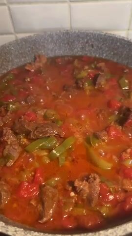The video shows a close-up shot of a large frying pan filled with a stew-like dish. The dish appears to contain chunks of beef, diced tomatoes, and sliced green peppers in a reddish-brown sauce. The stew is simmering, with bubbles appearing on the surface. The frying pan is a light grey color with a speckled texture. Behind the pan, white tiles are visible on the wall. The camera remains mostly still, with slight movements due to hand holding.
