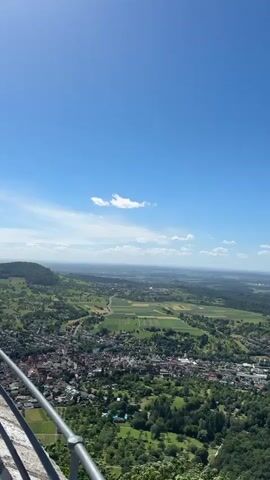 A vertical video captures a sweeping panoramic view of a rural landscape on a clear, sunny day. The sky is a vibrant blue with a few wispy white clouds. In the foreground, a metal railing is partially visible, suggesting the vantage point is from an elevated structure or viewpoint, possibly a balcony or a cliff edge. Below, a town is nestled amidst rolling green hills and lush forests. Houses with red-tiled roofs are clustered together, interspersed with trees and smaller patches of greenery. Fields with various shades of green and yellow are visible, indicating agricultural activity. In the middle distance, a large building complex with a white roof and what appears to be solar panels is situated amongst the trees. The landscape stretches out towards the horizon, where the terrain becomes flatter and the details are softened by the distance. The overall impression is one of natural beauty and tranquil rural life.