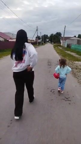 A woman and a young child walk away from the camera down a paved road. The woman is wearing a white sweatshirt with a graphic on the back and black pants. The child is wearing a light blue sweatshirt and blue jeans, and is carrying a red purse. The road is lined with houses and fences. The sky is cloudy.