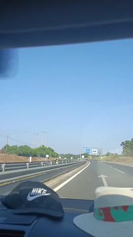 A video begins with a view from inside a car, looking out the windshield. A black Nike baseball cap sits on the dashboard in the foreground, and a white sun hat with a colorful band is partially visible to the right. The car is on a highway, with a clear blue sky overhead. Trees and utility poles line the side of the road, and there are guardrails separating the lanes. A road sign is visible in the distance. The camera pans slightly to the right, revealing more of the highway and the surrounding landscape. The video ends with a shot of the car's interior ceiling.