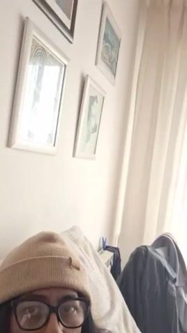 A woman wearing glasses and a tan beanie lies on a bed. She's wearing a black and white striped shirt. She sticks out her tongue, then puts a round cookie in her mouth and eats it. There are two framed pictures on the wall behind her, as well as curtains and some clothes hanging nearby.