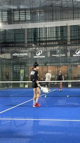 This video captures a group of people playing padel at an indoor court. The scene is set in Ban Buak Khrok North, Thailand. The court has a bright blue surface with white lines. A net divides the court in half. In the background, there are glass walls and a metal roof structure typical of an indoor sports facility.

The main subject is a woman in the foreground, wearing a black shirt with the word "MUSCLE" on the back and black shorts. She is holding a padel racket and appears to be in the middle of a game. Her body is turned away from the camera, suggesting she is reacting to a ball. She is wearing bright pink sneakers.

In the background, on the other side of the net, two other women are visible. One is wearing a white shirt and shorts, and the other is in a black top and shorts. They are also holding rackets. Another person, a man in a white shirt and beige shorts, is also on the court, further in the background. Several yellow padel balls are scattered on the court.

Visible text includes logos and names on the banners in the background. These include "Niko Padel Academy," "DECATHLON," and "Padel Club." The overall atmosphere appears to be active and engaged, indicating a sports match is in progress. The lighting suggests it could be daytime or a well-lit indoor facility.