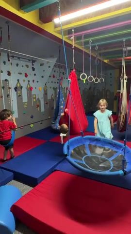 The video shows a brightly colored indoor play space. On the left, a young child wearing a red shirt and black shorts is on a zipline, swinging forward. In the center, a child is inside a large red fabric swing. To the right, a young girl in a light blue dress is running on a red mat and looking towards the camera. Another child is partially visible in the red swing. The room has a rock climbing wall with colorful holds and a cityscape mural on the back wall. Various swings and suspended equipment hang from the ceiling. The floor is covered in red and blue mats. A woman is standing on the right side of the frame, looking towards the children.