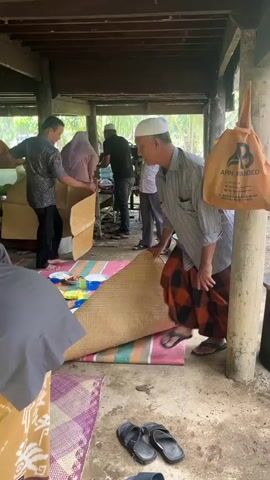 A group of men are gathered in an outdoor, covered area. Some are wearing white skullcaps and traditional Islamic clothing. One man is holding up a woven mat, showing it to another man who is facing away from the camera and is dressed in a grey shirt and traditional orange and black patterned sarong. Several other people, including women in headscarves, are visible in the background, along with tables set with food. The ground appears to be dirt and concrete, with mats laid out for seating. The environment suggests a communal gathering or meal.