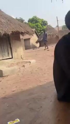 The video shows a man sitting at a wooden table in a rural area. In the background, there are several small, simple buildings with thatched roofs. Another man is walking in the distance between the buildings. The camera pans around to show different angles of the scene and the surrounding environment. The man sitting at the table is holding a mobile phone. The ground is dirt or sand. The sky is bright with some clouds.
