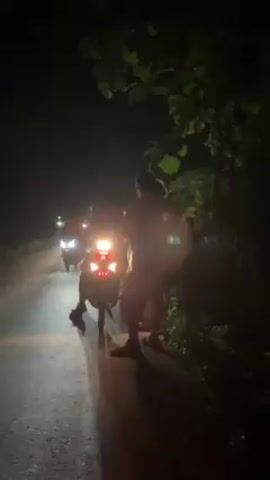 The scene takes place at night on a narrow paved road, bordered by trees and foliage on the right side. Several people stand near the edge of the road while others are on motorbikes with their headlights illuminated, facing towards the viewer. The light of the headlamps is casting light on the people standing and on the road. Some of the individuals are barefoot. The environment appears rural or suburban, given the presence of trees and the narrow road. The people seem to be interacting or observing something in the direction the motorbikes are facing. The overall impression is a nighttime gathering of people on a roadside.