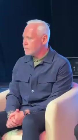 A man with short white hair and a small beard sits in a chair. He is wearing a navy blue shirt over a green collared shirt, dark pants, and multiple bracelets on his left wrist. He has his hands clasped in front of him. He appears to be listening intently. Behind him is a black background. The chair is a light tan color. The man's face is serious, and his expression is neutral. The lighting suggests a stage setting. There is audio of a male voice speaking.