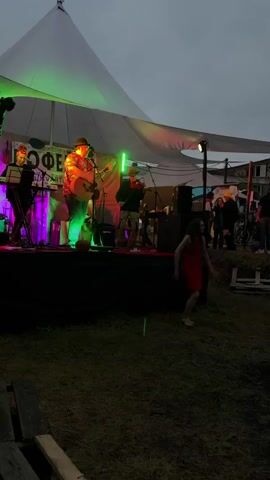 A live band is performing on a stage under a white tent. The band consists of a guitarist and singer in a colorful shirt, a keyboardist, and a violinist. The stage is lit with colorful lights, including purple and green. A young woman in a red dress is dancing in front of the stage. There are people in the background, some watching the performance, others milling about. The setting appears to be an outdoor event, possibly a festival or fair, with grass and some wooden structures visible. The sky is overcast.