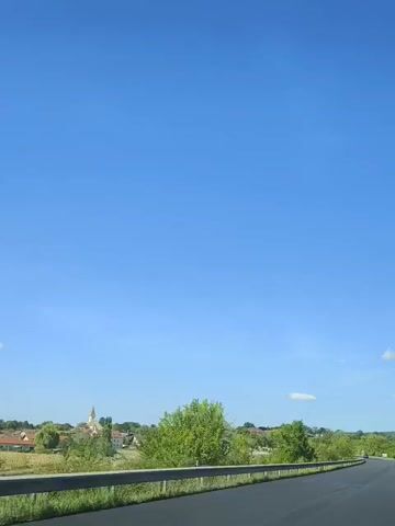 The video shows a view from a vehicle traveling on a road. The sky is clear and blue, with a few wispy clouds. On the left side of the road, there is a metal guardrail. Beyond the guardrail, there are green trees, bushes, and some buildings, including a church steeple. The road appears to be a highway or a main road, as it is paved and has a guardrail. The sun is shining, casting shadows and illuminating the scene. The overall impression is of a pleasant day, possibly in the countryside.
