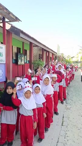 A line of children, mostly girls, dressed in red and white school uniforms and hijabs are standing outside a building. They are holding small white papers with red markings in their hands, raising them in the air. The children appear to be in an outdoor school setting, with a red and green building visible in the background. The weather appears to be sunny.