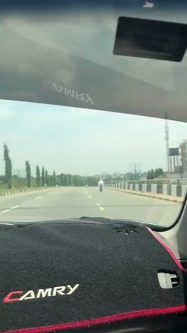 This video captures a first-person perspective from inside a car driving on a multi-lane highway in Aso, Nigeria, during daytime. The black dashboard of the car, covered by a mat displaying "CAMRY" in white text with a red border, is visible in the foreground. This text is also reflected inversely on the car's windshield. Ahead on the road, a person dressed in white clothing is riding a motorcycle, followed by other vehicles in the distance. The highway is lined with tall, slender trees and concrete barriers. In the background, a hazy sky extends over distant buildings, power lines, and billboards. The primary activity is driving, with accompanying audio of a person speaking in what sounds like Pidgin English, mentioning "salary this month," "next month," and "exam."