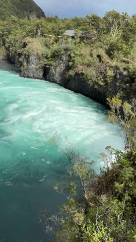 From a high vantage point, the camera offers a landscape view of a river, split in color and activity. The left side of the river is a deep, darker blue, flowing smoothly, while the right side presents a vibrant, bright turquoise, characterized by churning whitewater rapids. This contrast highlights the river's dynamic nature.

On either side of the river, steep banks rise, covered in lush greenery and trees that give way to forested hillsides. A small building is visible amidst the foliage on the right bank, suggesting human presence in this otherwise natural setting. The sky above is overcast, with dark clouds hinting at potentially stormy weather. The camera remains still, allowing the viewer to take in the full scope of the scenery and the river's powerful currents.