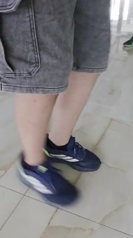 The video shows a person standing on a polished floor. The person is wearing blue sneakers, grey cargo shorts, and a light green t-shirt with a graphic design that reads "CHILL DAY." The person is in motion, appearing to be jumping or skipping. The environment appears to be an indoor space, possibly a warehouse or a large room, with reflections on the floor and shelving units visible in the background.