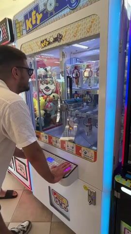A man wearing sunglasses and a white short-sleeved shirt plays a claw machine game. He is standing in front of the machine, with his hands on the controls. The claw machine is filled with stuffed animals and other prizes. The man is looking intently at the prizes inside the machine. The background shows a brightly lit arcade environment.