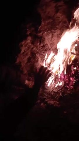 A person, possibly wearing a dark beanie and a dark-colored striped jacket, is warming their hands by a small campfire in the dark. The fire is relatively small but emits a bright white and red light. The ground around the fire appears to be covered with dry grass or leaves. The person is crouching down close to the fire.