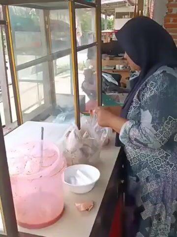 A person wearing a hijab and a patterned dress is standing behind a glass counter. They are holding plastic bags containing food items. To the left of the person is a pink dispenser with a straw. A white bowl is placed on the counter in front of the person. In the background, a person wearing a cap is visible reflected in the glass, along with a motorcycle and other objects. The environment appears to be an outdoor food stall or market.