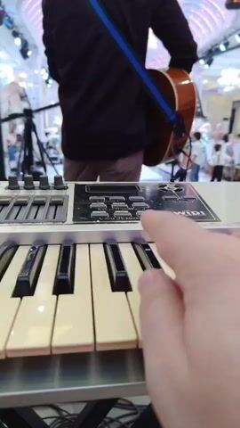 The video shows a first-person perspective of someone playing a keyboard. In the background, a band is performing in what appears to be a banquet hall or event space. One person is playing an acoustic guitar, and two others are seated at keyboards or electronic music production devices. The environment is decorated with lights and ornate architectural features. The camera is focused on the keyboard being played, with the hands of the player visible.