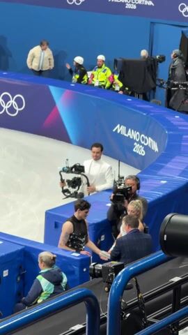 The video captures a scene at an ice rink, identifiable by the visible ice surface and the "MILANO CORTINA 2026" signage, likely indicating the upcoming Winter Olympics.  Several individuals are present, including two figure skaters dressed in performance attire, one in a black costume and the other in a silver sequined outfit. They are interacting with people who appear to be crew members or staff, possibly discussing something related to their performance. A camera operator is filming them, and another person in a white suit is holding a camera rig and surveying the scene. Other individuals, some wearing official-looking jackets and hard hats, are standing in the background, possibly representing event staff or security.  The environment includes blue barriers around the ice, cameras, and various pieces of equipment associated with video production.