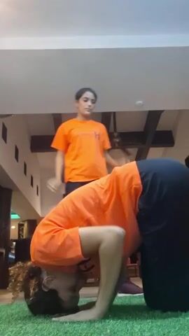 The video shows a person in an orange shirt and black pants performing a headstand on green artificial turf. They are supported by their hands and head, with their legs initially bent and then extended upwards. Another person, also in an orange shirt and black pants, stands behind them, seemingly providing support and encouragement. The scene takes place in a large indoor space, possibly a gym or studio, with high ceilings, visible structural beams, and some furniture in the background. The lighting appears to be artificial and diffused, creating a well-lit environment. A sign with a name on it is visible on the shirt of the standing person. The overall impression is that the video captures a fitness activity or yoga practice.