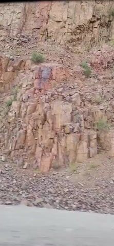 The video shows a rocky mountainside, likely a quarry, in a dry, arid environment. The rock faces are layered and fractured, with scree slopes of loose stones at the base. Sparse vegetation, mostly shrubs and grasses, grows in patches on the slopes and at the top of the mountain. The sky is a pale blue, suggesting it is daytime, possibly late afternoon given the lighting. The camera appears to be moving, possibly from a vehicle, as the view pans across the landscape. There are no people or man-made objects visible other than what might be the edge of a road in the foreground. The overall impression is one of a rugged, natural landscape.