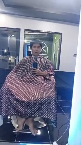 A person sits on a barber's chair, wearing a patterned cape. They hold a phone in their hand, appearing to take a selfie in a mirror. The person is wearing dark pants and their feet are bare. The environment looks like a barbershop, with mirrors and lighting visible. The person's facial expression suggests surprise or excitement.