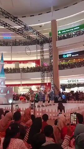 The video shows a performance taking place on a stage in the center of a shopping mall.  The stage is decorated with a white picket fence and a small castle prop. Several performers in blue and white costumes are roller skating on the stage. The audience, which includes people of different ages and ethnicities, are sitting or standing around the stage, many holding up their phones to record the performance.  The mall has multiple floors with several storefronts visible, including Crocs and Gripp. There are crowds of people watching from the upper levels. Overhead, a metal structure with a large hoop hanging from it is visible. The lighting is bright, and the atmosphere appears lively and crowded.