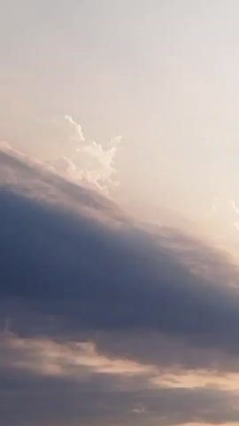 The video shows a time-lapse of a sunset. Dark clouds are visible in the foreground, with lighter, wispy clouds above. The sun, a small bright orb, emerges from behind the dark clouds and then disappears again. Silhouetted trees are visible at the bottom of the frame.