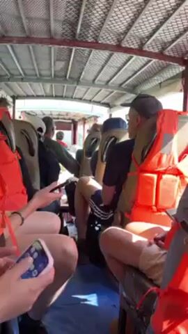 The video shows the interior of a boat with rows of seated passengers wearing orange life vests. Many are looking at their phones. The boat is moving through a river or body of water with green, tree-covered mountains on the side. The sky is clear and blue. The boat's wake is visible.