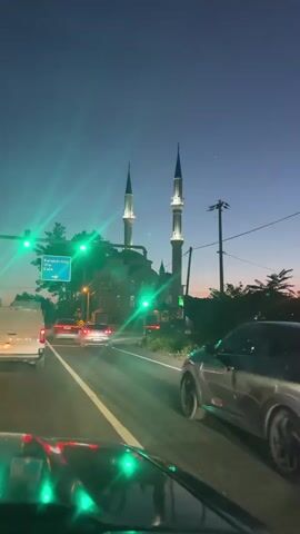 The video shows a car approaching a traffic light at an intersection. The light is green. In front of the car, traffic is also moving forward. Visible in the background is a mosque with two tall minarets. There is a sign pointing towards Karabörtlen, Ula, and Kale. The sky is darkening, indicating either early morning or late evening. The video is shot from the driver's perspective, and ambient music can be heard.