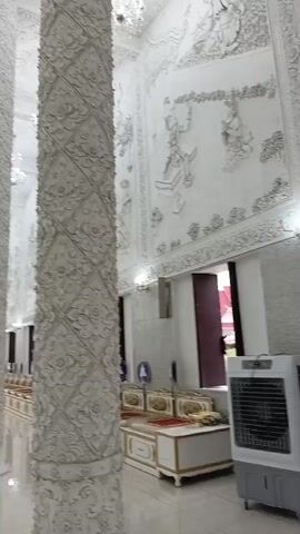 The video pans across the interior of a large, ornate building, possibly a temple or a church. The dominant color scheme is white, with intricate white carvings adorning the walls, columns, and ceiling. Several tall, elaborately carved white columns stand in the foreground, extending towards a high, decorated ceiling from which crystal chandeliers hang. The walls are decorated with bas-relief carvings of figures and patterns. In the background, at the far end of the hall, a large white statue, likely of a deity, is visible, surrounded by offerings. The floor is highly reflective, mirroring the white decor and the hanging lights. A person is seen walking in the distance towards the statue. In the right foreground, a portable air cooler is visible. The overall impression is one of grandeur and religious solemnity.
