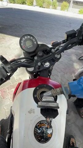The video shows a person fueling a motorcycle at a gas station. The motorcycle is white and red with a sticker on the fuel tank. A blue fuel nozzle is inserted into the motorcycle's fuel tank. In the background, a gas pump with "Indian Oil" branding is visible, along with other vehicles and a person wearing a mask and holding a fuel hose. The environment appears to be outdoors with a clear blue sky and mountains in the distance. The person filming is wearing a helmet and gloves.