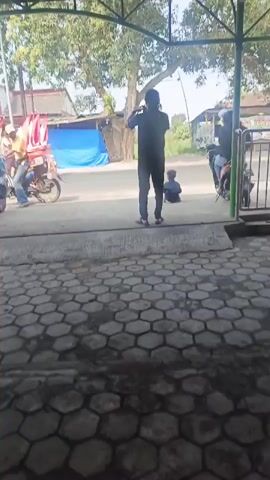 The video shows a group of people marching outdoors, holding Indonesian flags. Some individuals are on motorcycles, and there are trees and buildings in the background. The ground is paved with interlocking bricks. One person in the foreground is standing and facing away from the camera. Another person is sitting on the ground further back. The lighting suggests it is daytime.