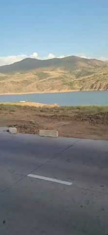 The video shows a landscape with a road in the foreground and a body of water with mountains in the background. The sun is shining and the sky is blue. The road appears to be asphalt with white lines marking the lanes. There are concrete barriers along the side of the road. The land between the road and the water is dry and covered with dirt and some vegetation. The water is calm and reflects the sky and mountains. The mountains are rugged and appear to be dry and covered with sparse vegetation. The overall scene is peaceful and scenic.