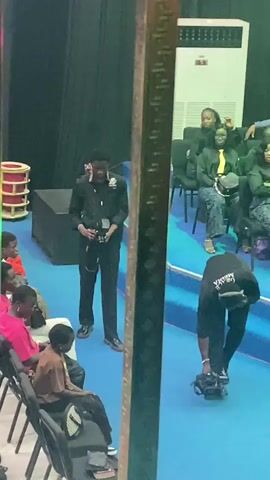 The video shows an indoor scene, possibly a stage or event space, with blue flooring and dark draped curtains in the background. In the foreground, a metal vertical beam partially obstructs the view.

On the left side of the frame, several people are seated in chairs facing towards the right. One person in a pink shirt is clearly visible. In the center, a person in a black outfit, possibly a uniform with a badge, stands holding a camera or similar device.

On the right side of the frame, a person in a black shirt with white lettering on the back, bending over to adjust a video camera on a tripod. Several other individuals are seated on raised blue tiered seating in the background, some wearing black tops and yellow accents. A large white air conditioning unit is visible on the far right in the background. The lighting appears to be artificial.