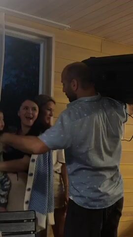 A man with a shaved head and a light blue patterned short-sleeved shirt is carrying a large black speaker on his right shoulder. He is facing away from the camera, looking towards a woman and a young child. The woman is holding the child, who is wearing a light green long-sleeved shirt and camouflage pants. The woman is wearing a light-colored cardigan over a dark top. The man is holding a smartphone in his right hand, near his face, as if taking a picture or recording. The background appears to be an outdoor covered area with wooden paneled walls. There is a window on the left side of the frame showing darkness outside.