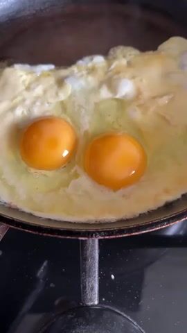 Two eggs are frying in a black pan on a dark stovetop. The egg whites are bubbling and solidifying, while the two bright orange yolks remain mostly intact in the center of the pan. The pan appears to be a bit used. The camera is positioned directly above the pan, offering a close-up view of the cooking process.