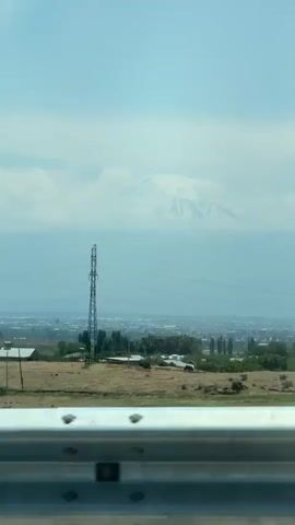 The media file captures a scenic view from a vehicle, likely on a highway given the reflective barrier in the foreground. The primary subject is a majestic, snow-capped mountain range dominating the distant horizon, partially obscured by a haze of clouds. Below the mountains, a sprawling city or town is visible, characterized by a dense cluster of buildings and structures. In the mid-ground, a tall, metallic power line tower stands prominently, with electric cables stretching across the frame. The foreground features a rural landscape with scattered houses, trees, and dry, grassy fields, suggesting a somewhat arid climate. The time of day appears to be daytime, with a bright, possibly slightly overcast sky. The overall mood conveyed is one of vastness and natural beauty juxtaposed with human development. There is no visible text in the media file. The location context provided is Voskehat, Armenia, which aligns with the visual cues of a mountainous landscape and a populated area.