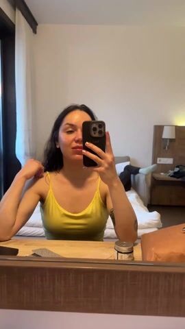 A woman is taking a selfie in a mirror in a hotel room. She is wearing a yellow tank top and has dark hair. She is holding a smartphone in her right hand and smiling. In the background, there is a bed with someone lying on it. There is also a nightstand with a lamp and some personal items. A jar and a brown bag are sitting on the shelf in front of the mirror.
