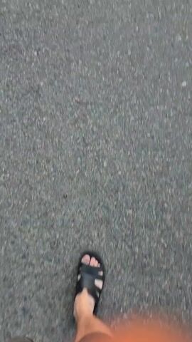 The video shows a first-person perspective of someone walking on an asphalt surface. The person is wearing black sandals and appears to be wearing shorts. The movement is slightly blurred, indicating the person is in motion. The lighting is subdued, suggesting it could be overcast or late in the day. The asphalt surface appears to be a path or road.