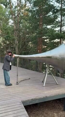 A person stands on a wooden deck near a large, silver, cone-shaped object that resembles a horn or megaphone. The person is wearing a dark jacket, blue jeans, and sunglasses, and they have their mouth to the narrow end of the object. The object is supported by two metallic stands. The background consists of trees and foliage, suggesting an outdoor, natural setting. The person appears to be speaking or making a sound into the object.